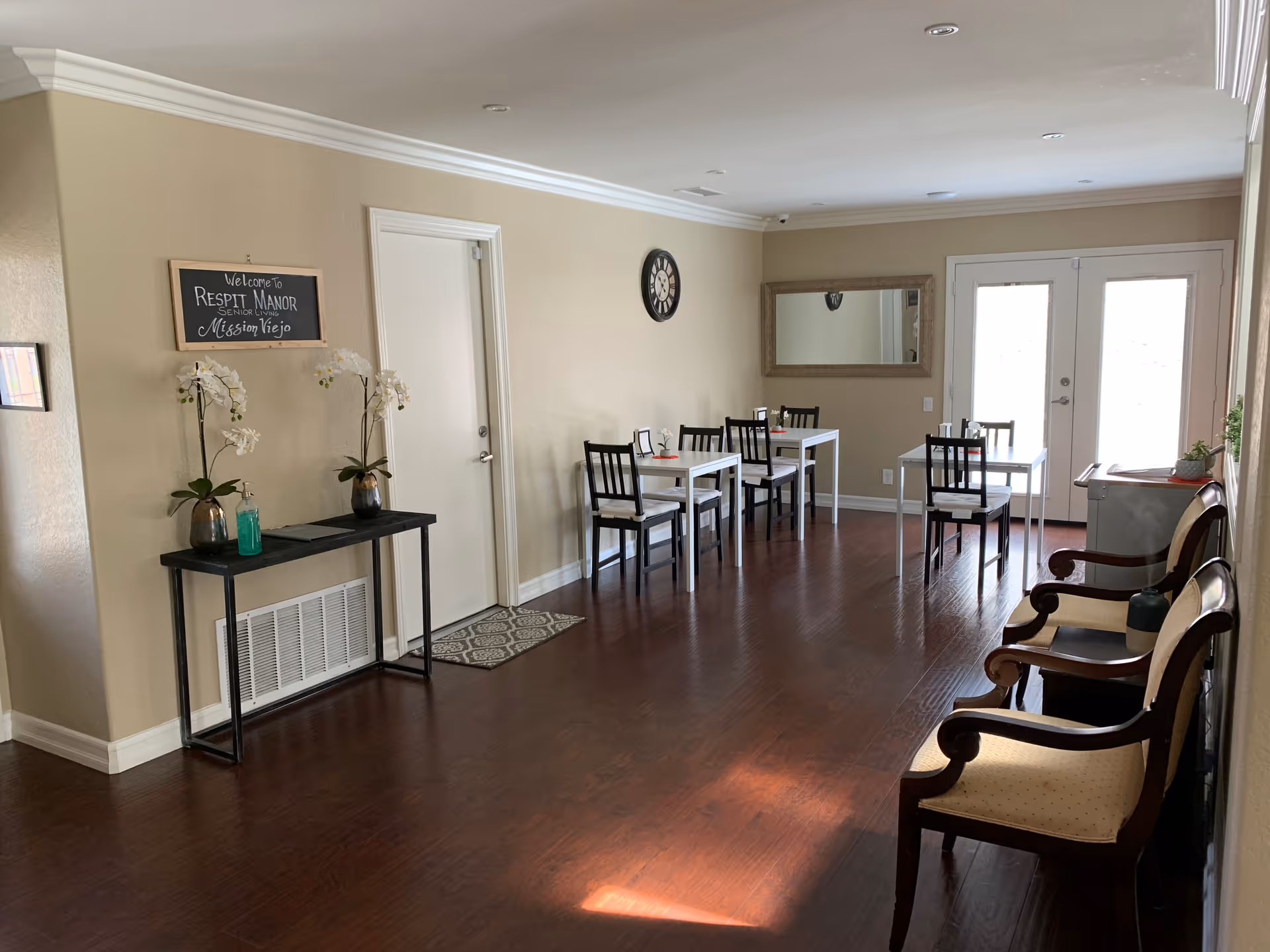 A bright room with wooden flooring featuring a small dining area with three white tables and black chairs. On the right side, there are three wooden armchairs with beige cushions. A black console table with two potted white orchids and a green bottle is against the left wall. Above the table is a chalkboard sign that reads 'Welcome to Respit Manor Senior Living Mission Viejo'. A large mirror and a clock are mounted on the far wall, and double glass doors allow natural light to enter the room.