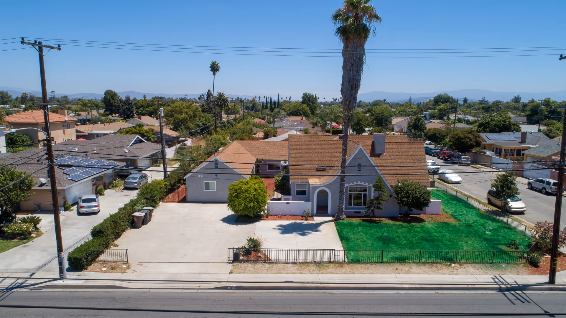 Aerial view of a residential neighborhood featuring a single-story house with a brown roof, a tall palm tree in the front yard, a green lawn, and a concrete driveway. Surrounding houses and trees are visible under a clear blue sky.