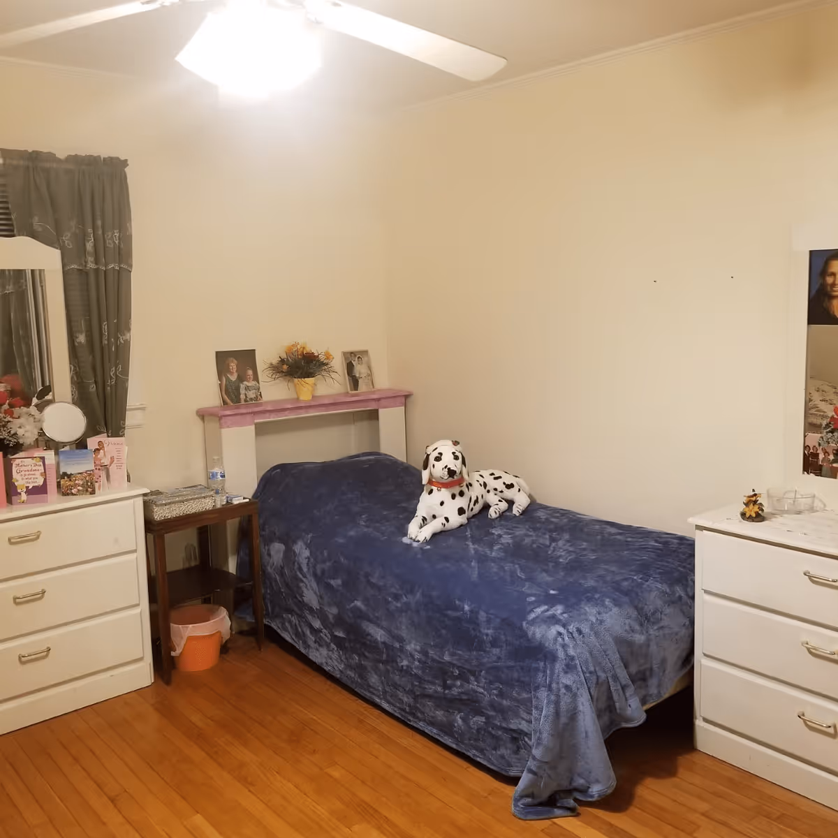 A small bedroom with a single bed covered in a blue blanket and a stuffed Dalmatian dog on top. The room has wooden floors, two white dressers with mirrors, a small wooden side table, and a window with green curtains. There are framed photos and decorative items on the dressers and a small flower arrangement on a shelf above the bed.