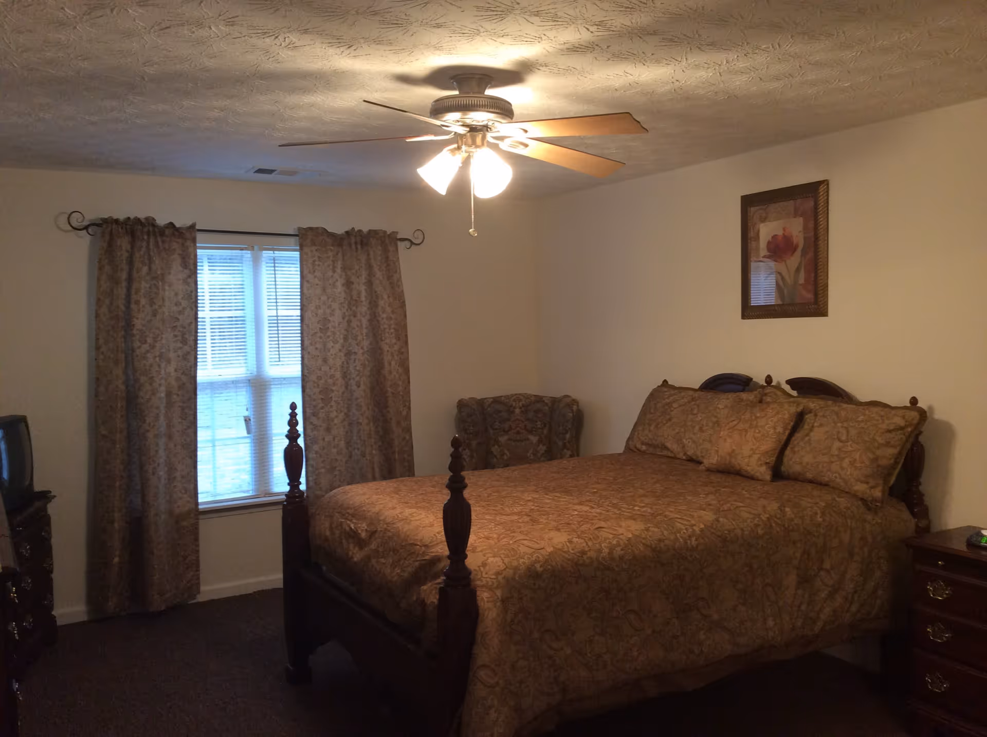 A bedroom with a large wooden bed covered in a brown patterned bedspread and matching pillows. There is a window with floral curtains, a ceiling fan with lights, a framed floral picture on the wall, a cushioned armchair in the corner, and a wooden dresser with a small TV on it.