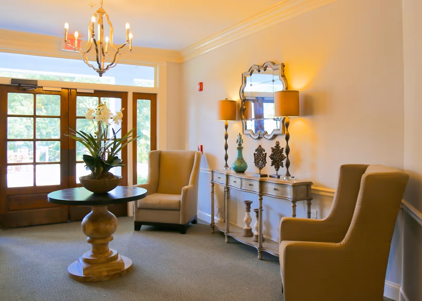A bright and welcoming sitting area with two beige armchairs, a round wooden table with a potted plant in the center, a decorative console table with lamps and ornaments, and a large windowed door letting in natural light.