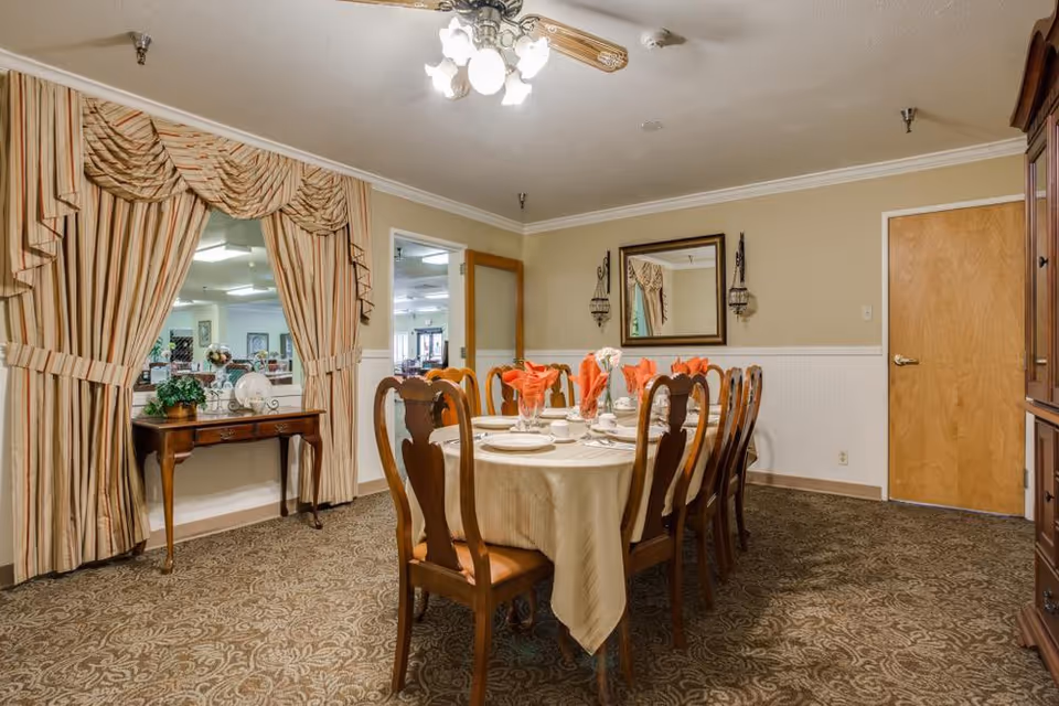 A formal dining room with a long table covered with a beige tablecloth, set with plates, cups, and orange folded napkins. The room has patterned carpet, beige walls with white wainscoting, a ceiling fan with lights, a large mirror on the wall, and a wooden sideboard with decorative items. There is a doorway with striped curtains leading to another room.