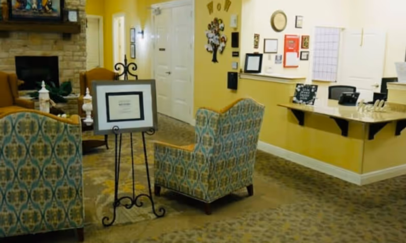 A cozy interior common area with patterned armchairs, a stone fireplace, a small table with decorative items, and a reception desk with various notices and a calendar on the wall behind it. The walls are painted yellow and the floor is carpeted.