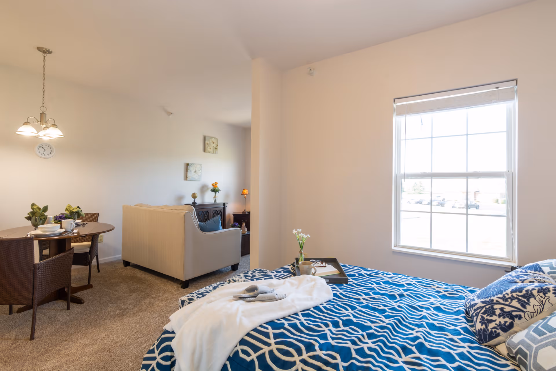 A bright and cozy studio apartment featuring a bed with blue and white patterned bedding near a large window, a small dining table with four chairs, and a beige sofa facing a TV stand with decorative items. The room is carpeted and softly lit with wall art and a hanging light fixture above the dining area.