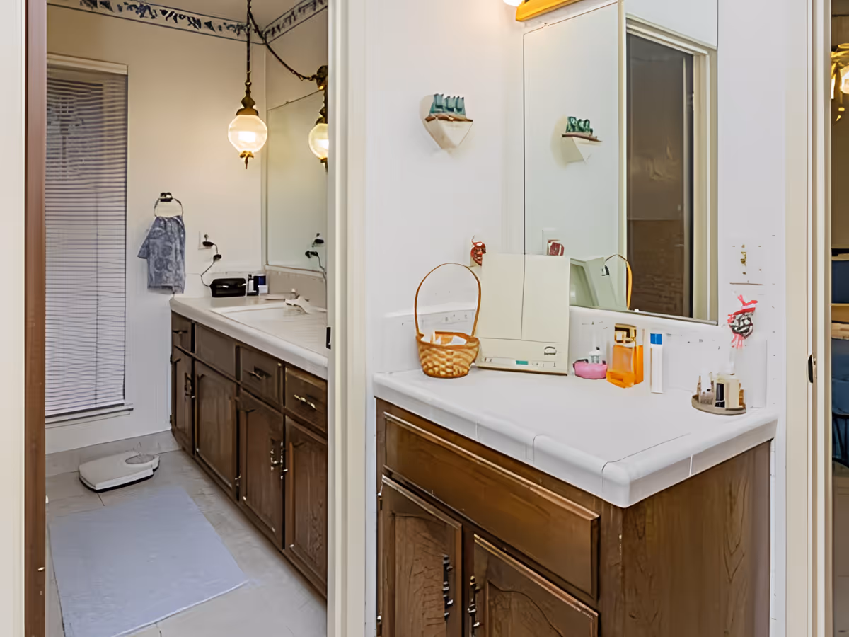 A bathroom with a long countertop featuring two sinks, wooden cabinets underneath, a large mirror above, and various toiletries on the counter. There is a hanging light fixture, a towel on a ring holder, a white bath mat on the floor, and a scale near a window with blinds.