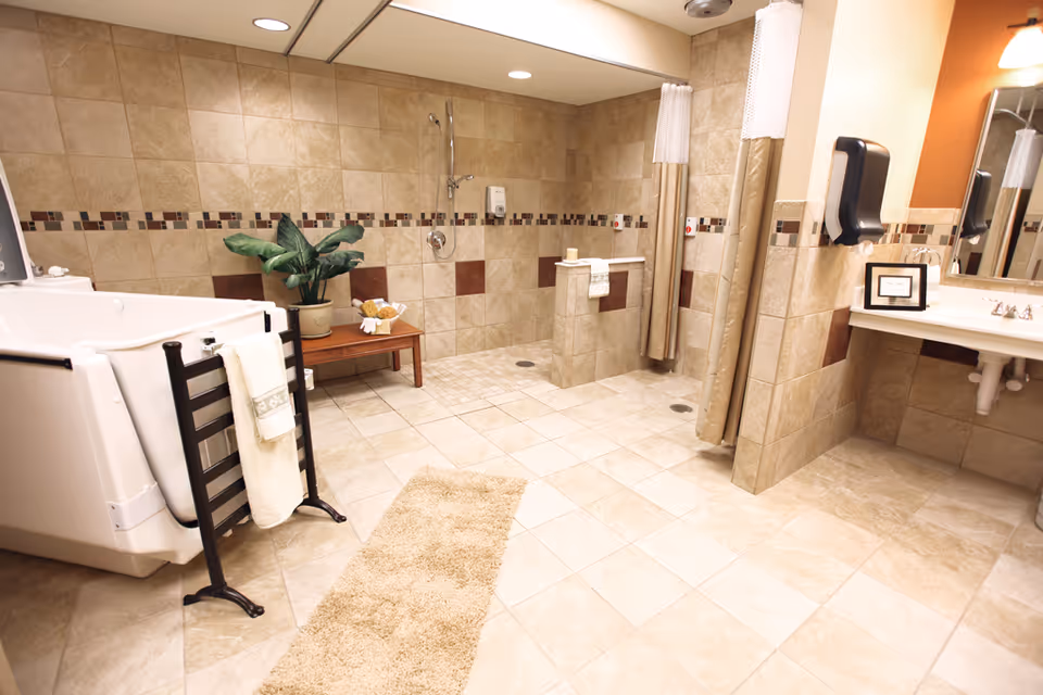 A spacious, well-lit bathroom with beige tiled walls and floors. The room features a large walk-in bathtub with a towel rack beside it, a wooden bench with a potted plant and towels, two shower stalls with curtains, and a sink with a mirror and soap dispenser. The decor includes a beige rug on the floor and an orange accent wall above the sink area.