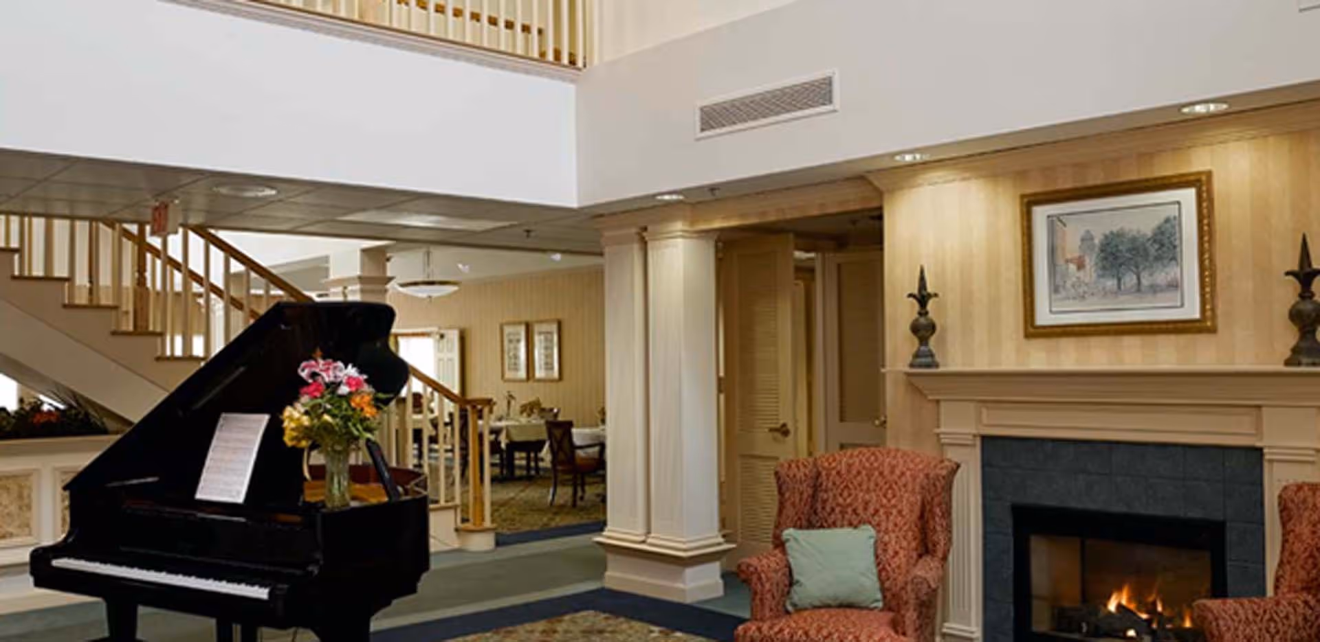 A cozy interior living area featuring a black grand piano with a vase of colorful flowers on top, a lit fireplace with a framed picture above it, and two patterned armchairs with cushions. In the background, there is a staircase with wooden railings and a dining area with tables and chairs.