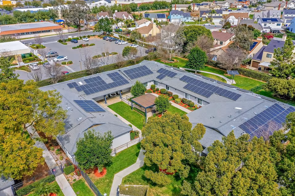 Aerial view of a senior living facility named Cottages at Riverside featuring a U-shaped building with solar panels on the roof, surrounded by green lawns, trees, and a parking lot with several cars. The facility is located in a residential neighborhood with houses visible in the background.