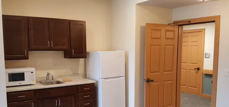 Interior view of a small kitchen area with dark wooden cabinets, a white microwave on the countertop, a stainless steel sink, and a white refrigerator. There is an open wooden door leading to a hallway with another closed wooden door visible.