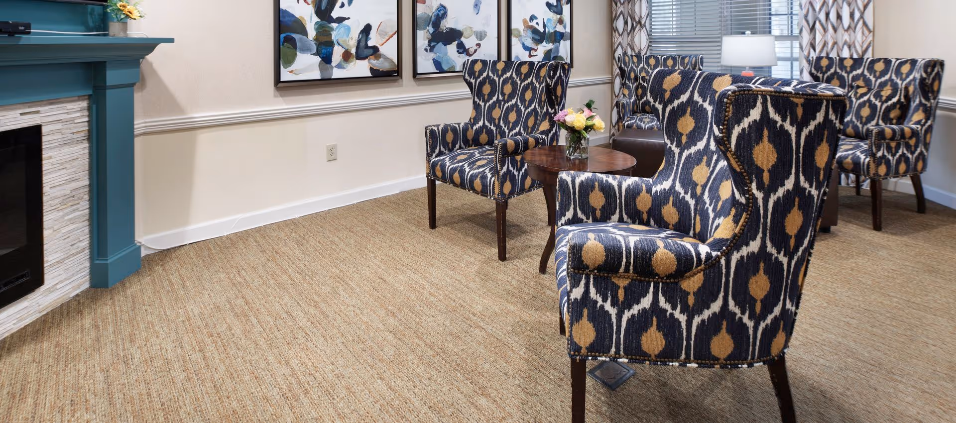 A cozy living room area with patterned armchairs arranged around a small wooden table holding a vase of flowers. The room features a teal fireplace on the left, abstract artwork on the wall, and a window with patterned curtains and a table lamp.