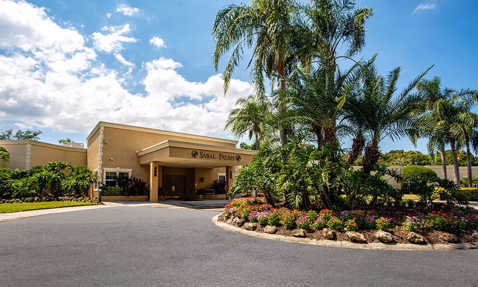 Front exterior view of Sabal Palms Health & Rehabilitation building with a driveway, landscaped garden featuring palm trees and colorful flowers, under a partly cloudy blue sky.