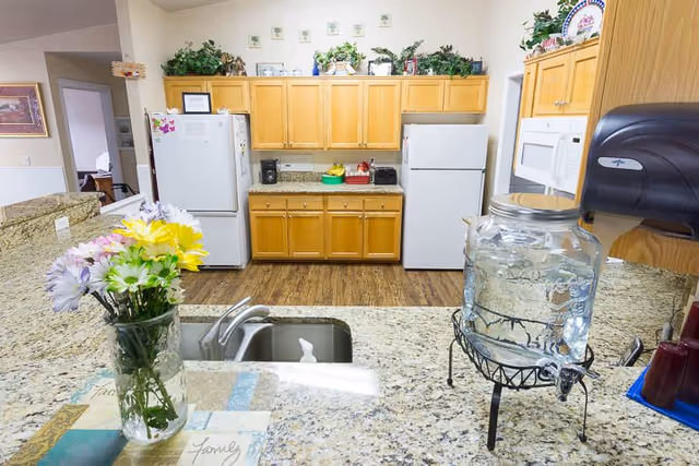 Community kitchen with granite countertops, sink, glass water dispenser and vase of flowers in foreground and wooden cabinets, microwave and two refrigerators in the background.