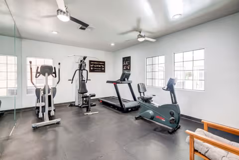 A small gym room with exercise equipment including an elliptical machine, a multi-function weight machine, a treadmill, and a stationary bike. The room has white walls, large windows letting in natural light, ceiling fans, and a cushioned wooden chair in the corner. A sign on the wall reads 'Gramercy Parc Gym'.