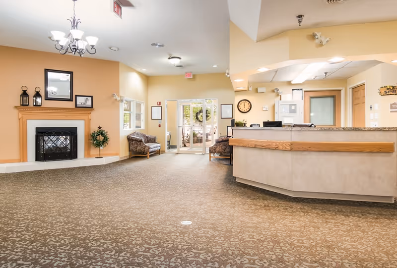 A spacious senior living facility lobby with a patterned carpet, a wooden reception desk on the right, two armchairs near the glass exit doors at the back, a fireplace with a wooden mantle on the left wall, and a chandelier hanging from the ceiling.