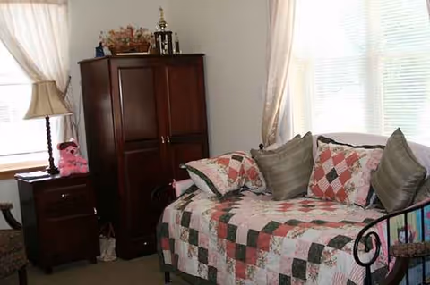A cozy bedroom featuring a patchwork quilt bed, tall dresser, nightstand, pink teddy, and sheer curtains.