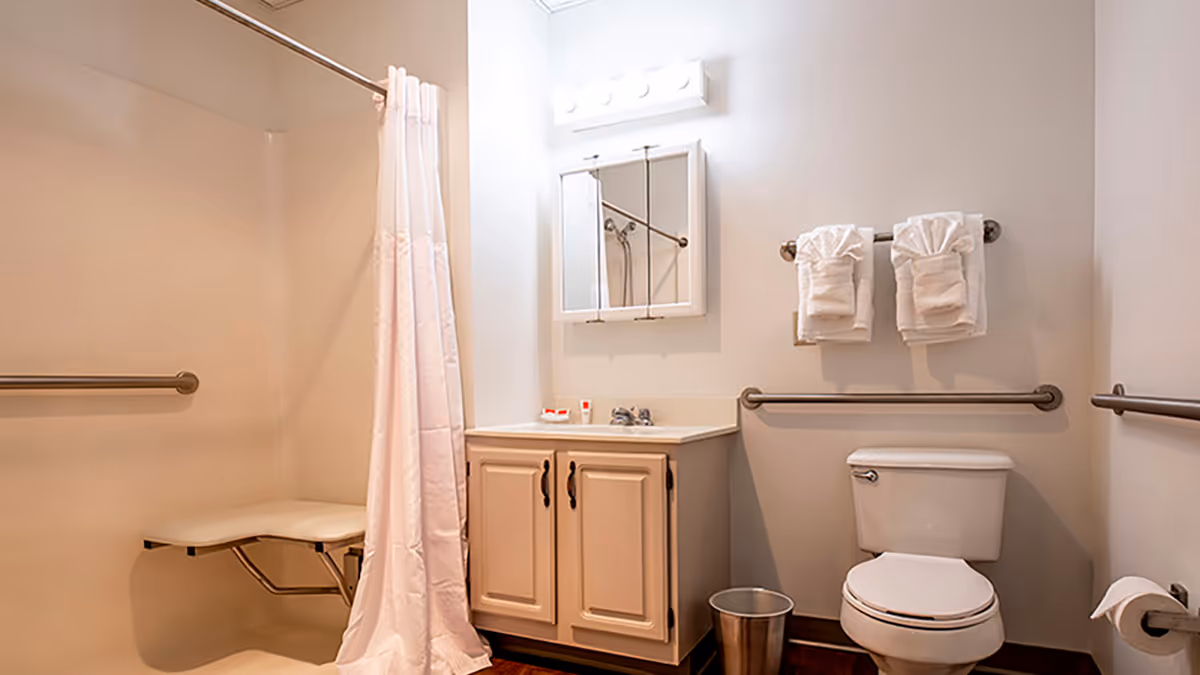 A clean and accessible bathroom featuring a shower area with a fold-down seat and grab bars, a white shower curtain, a vanity with a sink and mirrored medicine cabinet above, two neatly folded white towels hanging on a towel rack, a toilet with grab bars on both sides, and a small trash can on the floor.