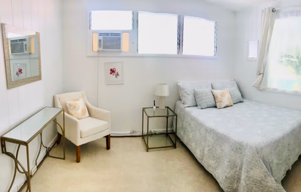 A bright and clean bedroom with a large bed covered in light gray bedding and several pillows. Next to the bed is a glass-top side table with a lamp and a small item on it. There is a white upholstered armchair with a decorative pillow, a glass-top console table, a wall mirror, and a framed flower picture on the wall. Three windows with air conditioning units are above the bed, and a curtained window is on the right side, letting in natural light.
