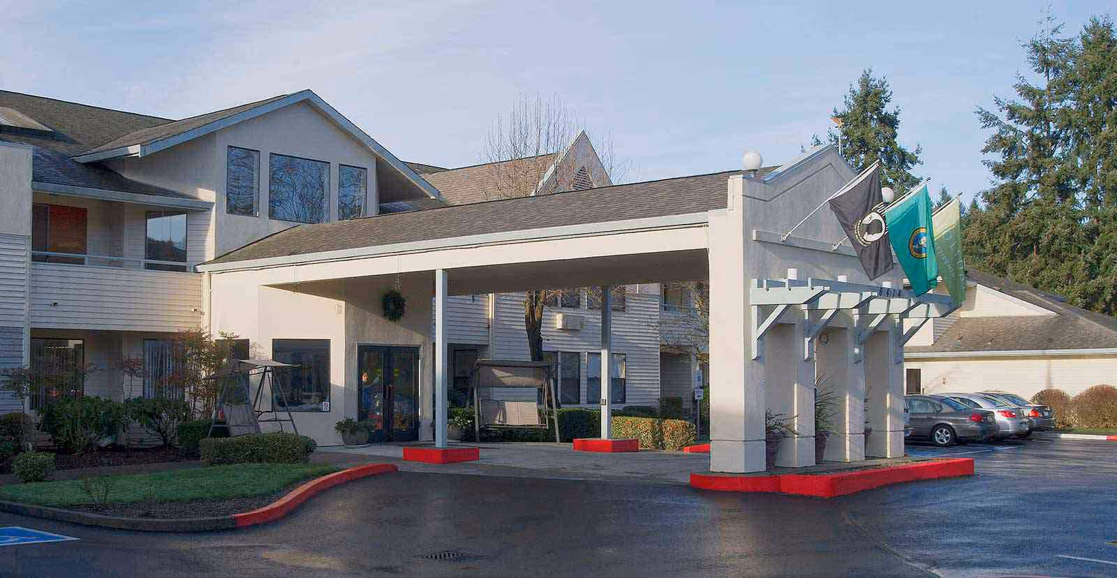 Exterior entrance of a senior living building with a covered porte-cochère, flags, and parked cars.