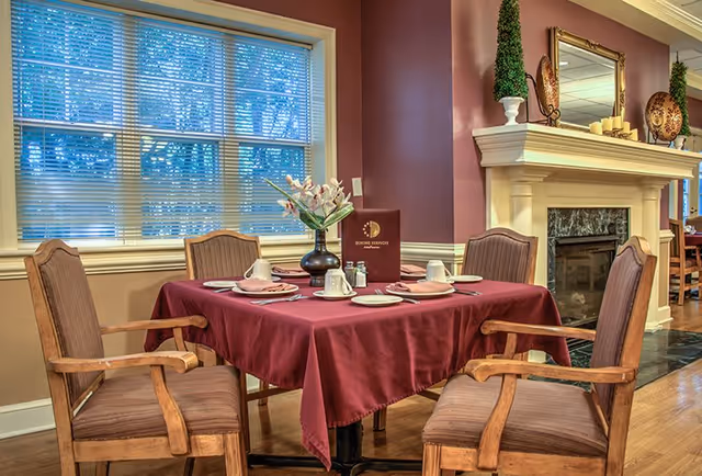A dining table set for four with a burgundy tablecloth, white cups, plates, and silverware. A vase with flowers and a dining services menu are placed in the center. The room has wooden chairs with cushions, a large window with blinds, and a fireplace with decorative items on the mantel.