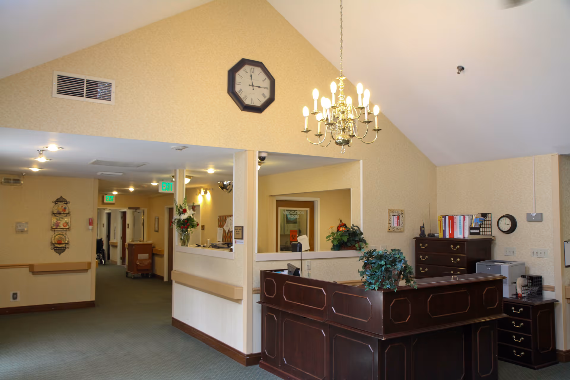 Interior view of a senior living facility reception area with a dark wooden front desk, a chandelier hanging from a vaulted ceiling, a wall clock, and a hallway leading to other rooms. The walls are light-colored with a subtle pattern, and there are plants and office supplies on the desk.