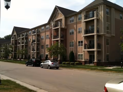 Four-story residential building with balconies and parked cars along a tree-lined street.
