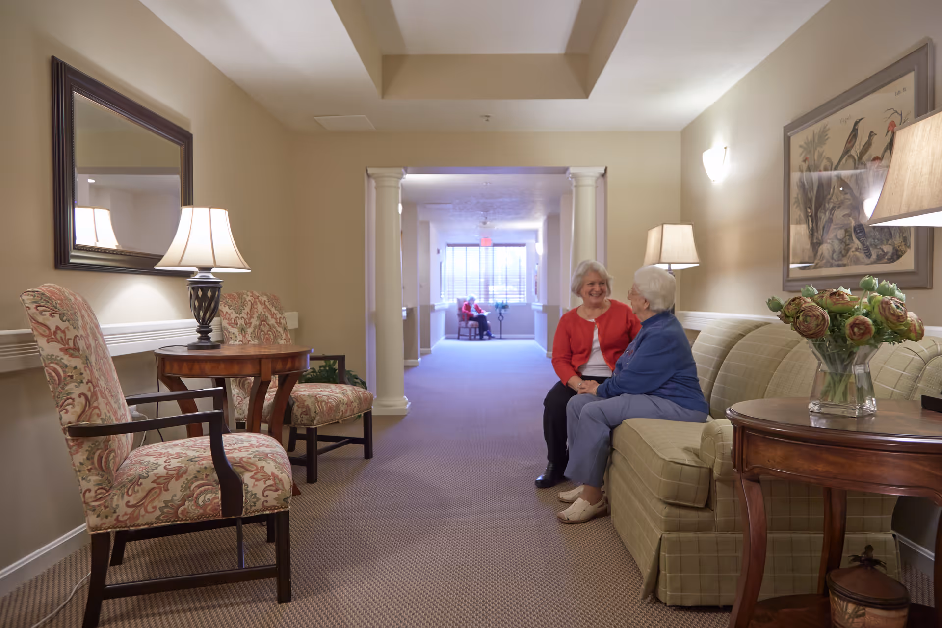 Two elderly women sitting and chatting on a green plaid sofa in a well-lit hallway lounge area with beige walls, floral armchairs, wooden side tables with lamps, a large mirror, and a framed bird artwork. In the background, another person is seated near a window at the end of the hallway.