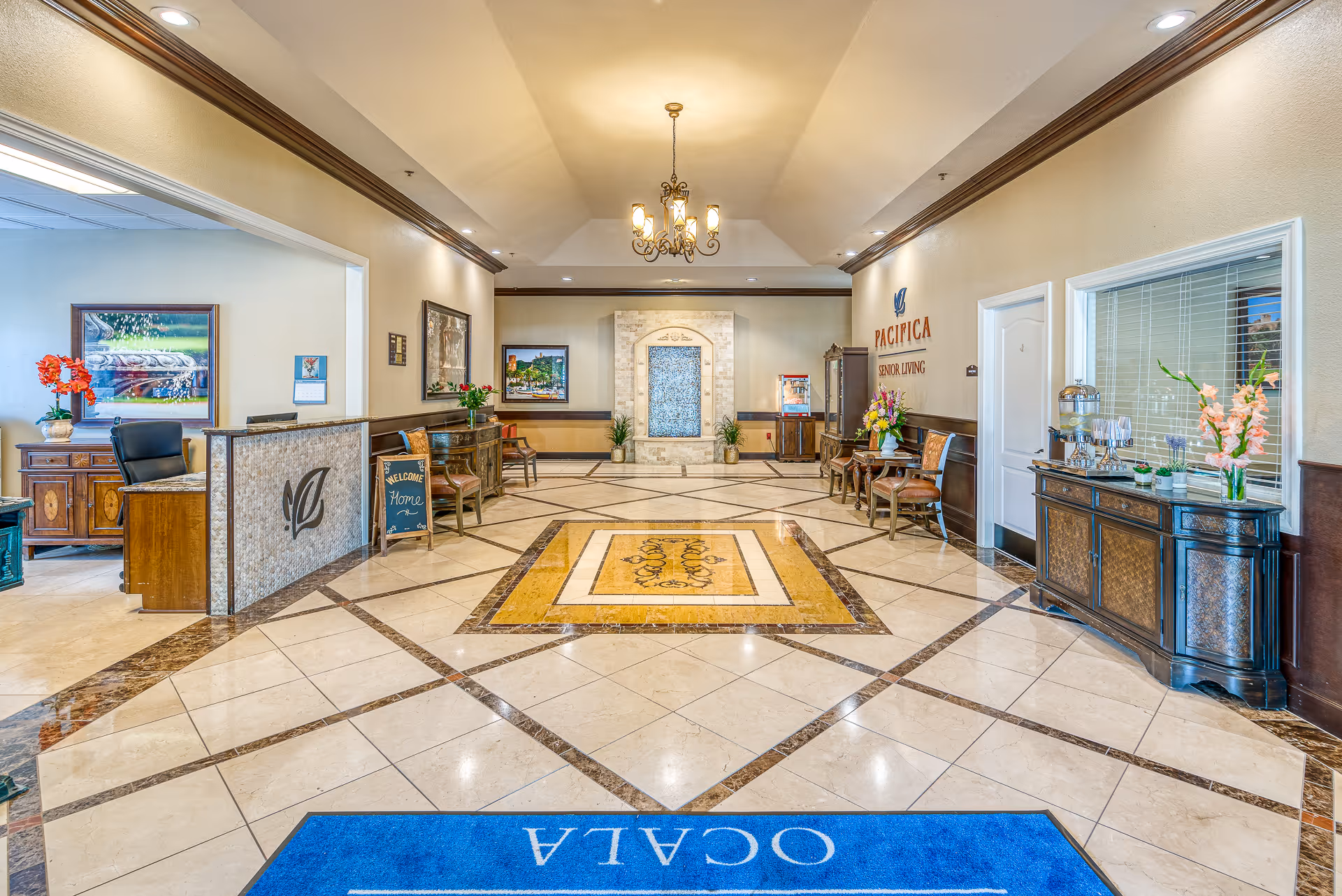 Bright and spacious lobby area of Pacifica Senior Living Ocala featuring a decorative tiled floor with a central yellow and brown pattern, a chandelier hanging from the ceiling, a water fountain against the far wall, several chairs and tables with flower arrangements, a reception desk on the left, and a blue welcome mat on the floor with the word 'OCALA'.