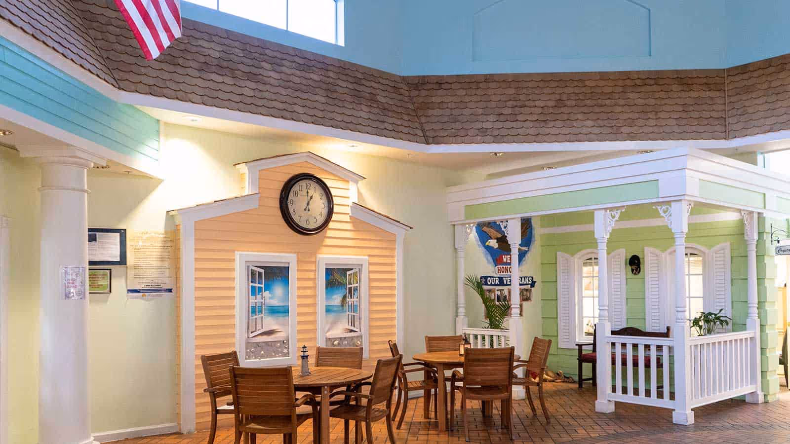 Indoor seating area in a senior living facility featuring two round wooden tables with chairs. The space is decorated with small house-like structures painted in pastel colors, one yellow with a clock and beach scene windows, and another green with white trim and a bench. An American flag hangs from the ceiling, and there is a sign honoring veterans on the wall.