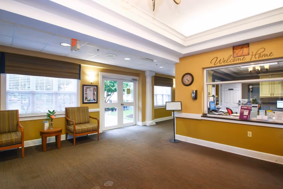Reception area of a senior living facility with a welcome desk on the right, a clock on the wall, and a sign above the desk that says 'Welcome Home'. There are two striped armchairs and a small wooden table with a plant on the left near large windows with blinds. Double glass doors lead outside, and the walls are painted yellow with white trim.