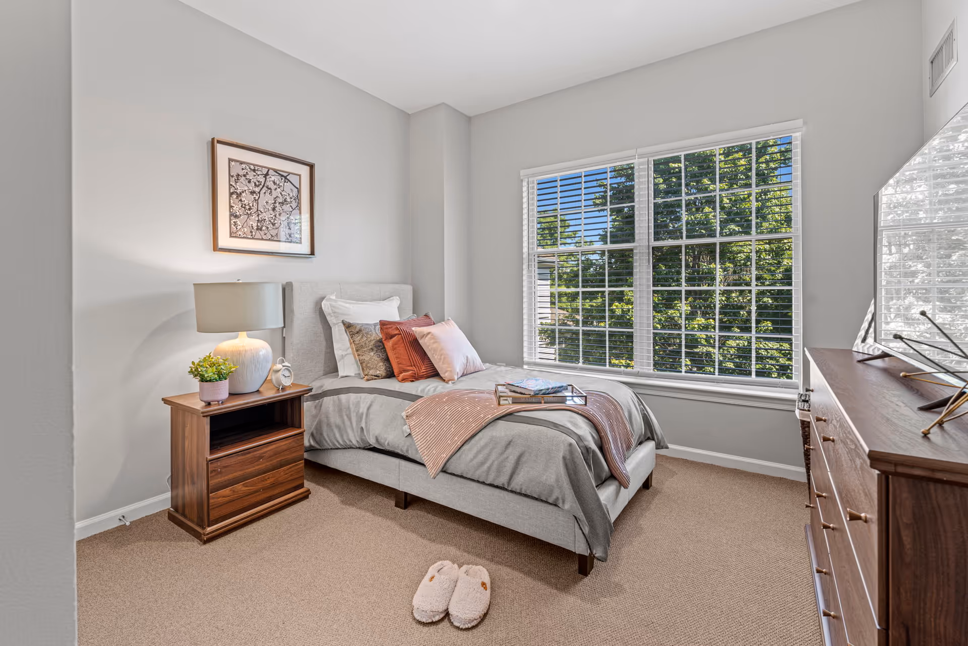 A bright furnished bedroom with a single bed, nightstand, dresser, large window and slippers on the carpet.
