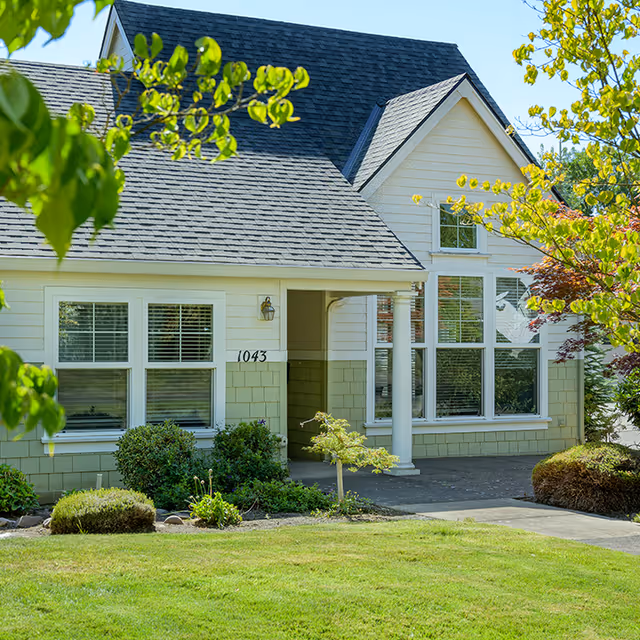 Exterior view of a single-story residential building with light green siding and white trim, featuring a covered entrance supported by a white column, multiple windows with blinds, and a well-maintained front yard with green grass and shrubs.