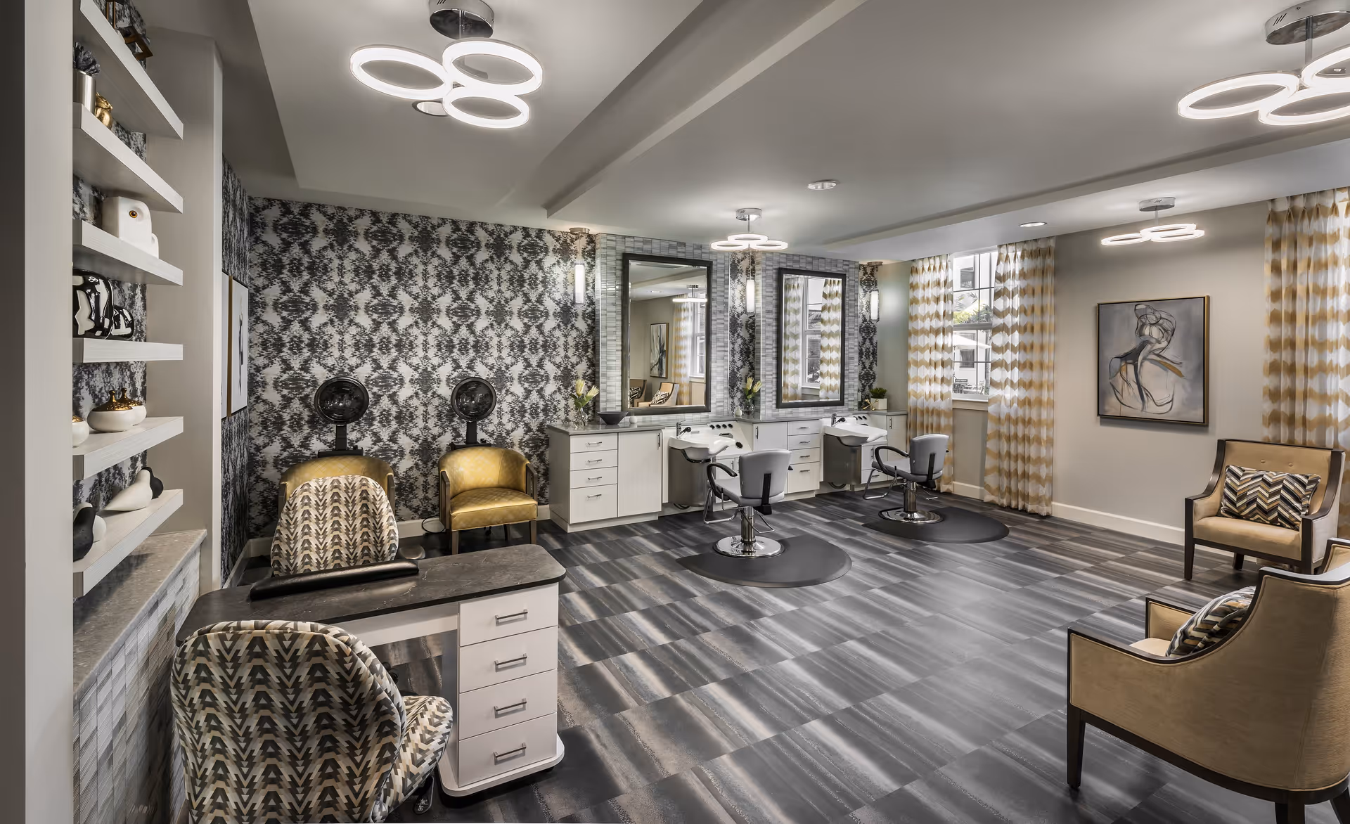 A modern hair salon area inside an assisted living facility featuring two hair washing stations with mirrors and chairs, a manicure table with patterned chairs, two yellow armchairs under hair dryers, and additional seating with beige armchairs. The room has patterned wallpaper, large windows with yellow and white curtains, contemporary circular ceiling lights, and decorative artwork on the walls.