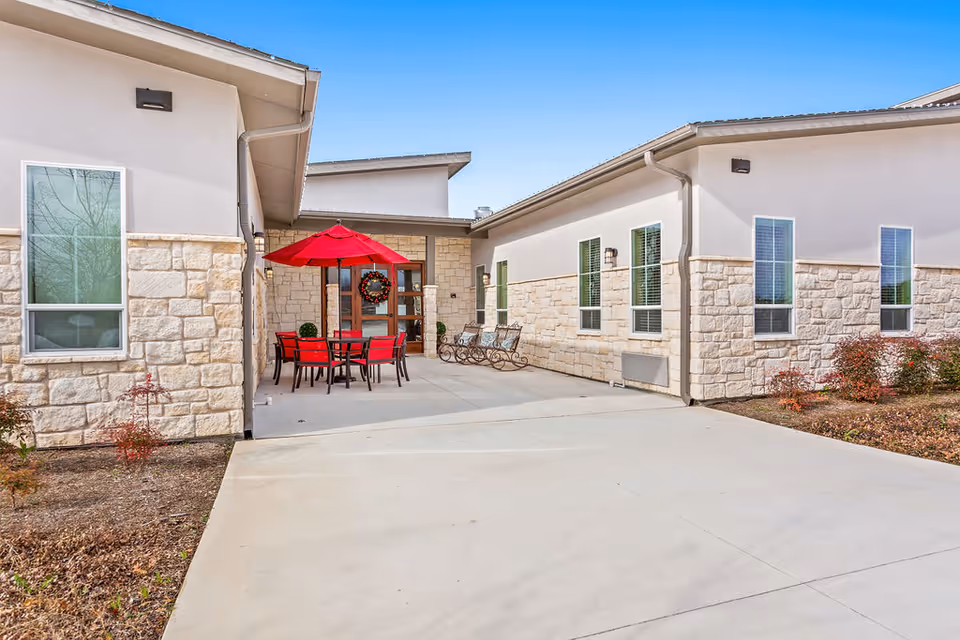 Outdoor courtyard between single-story building wings featuring a red patio umbrella, table and chairs in front of glass doors.