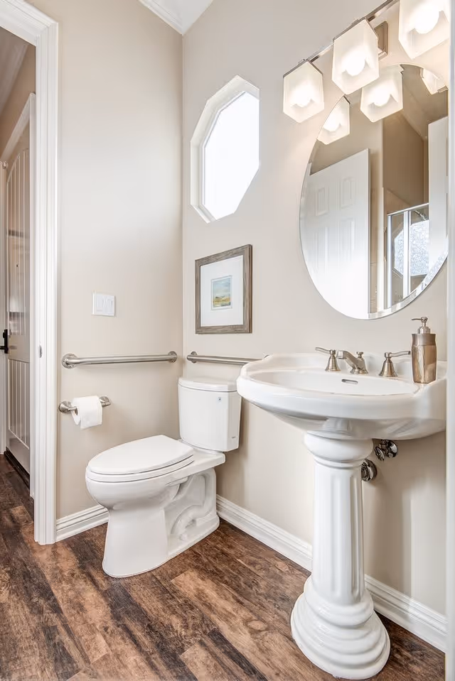 A clean and bright bathroom with a white pedestal sink, a round mirror above it, a toilet with grab bars on the walls, a small framed picture, and a window with frosted glass. The floor has a wood-like finish.