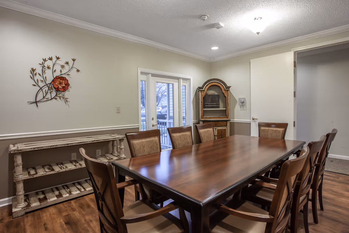 A dining room with a long wooden table surrounded by eight chairs. The room has light-colored walls with a decorative metal wall art featuring a red flower. There is a rustic wooden shelf against one wall and a vintage cabinet in the corner. Double glass doors lead outside, and the floor is wooden.