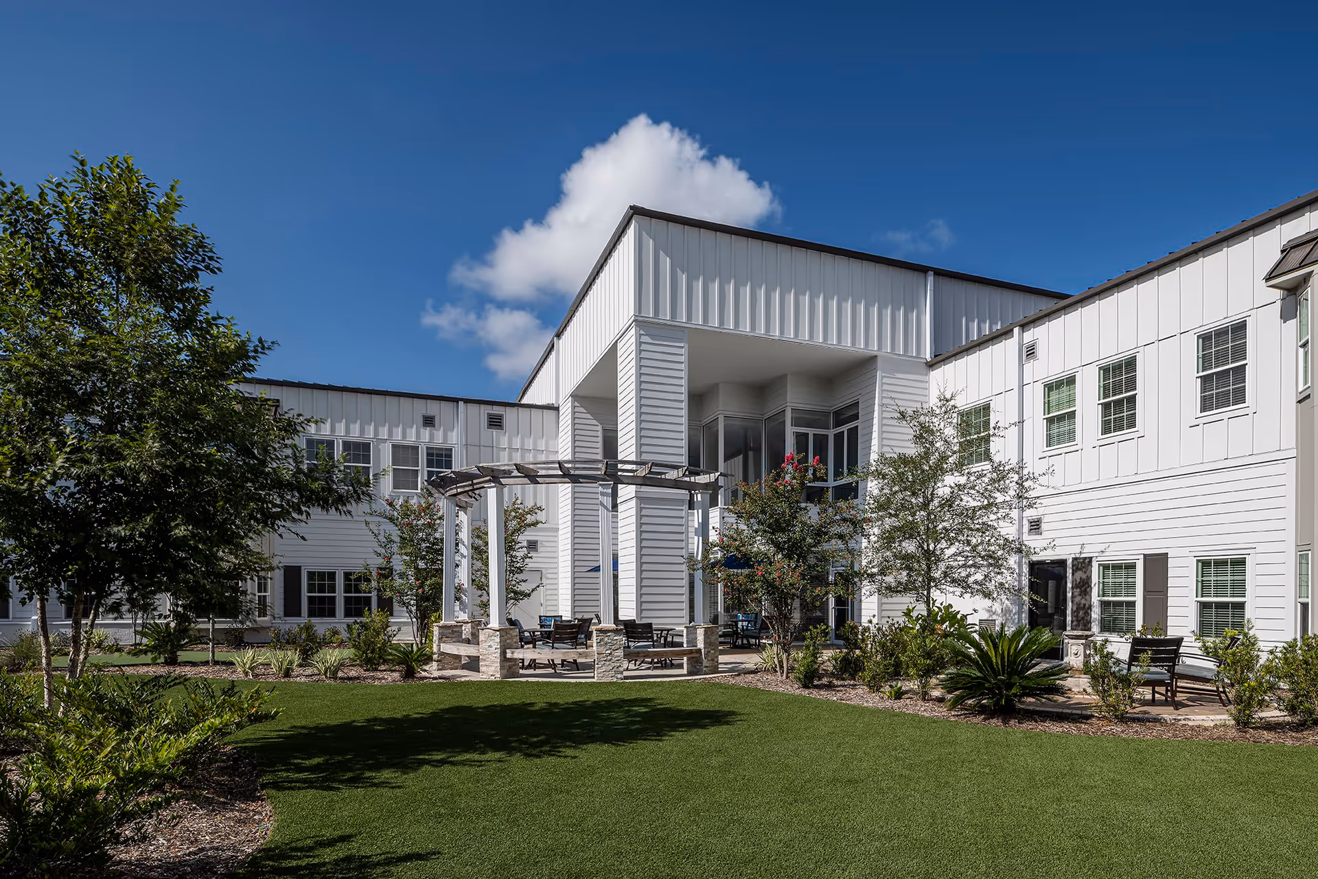 Outdoor courtyard area of a senior living facility with a white pergola, seating, green lawn, trees, and shrubs under a blue sky with some clouds. The building surrounding the courtyard is white with multiple windows.