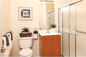 A clean bathroom featuring a toilet, a wooden vanity with a white countertop and sink, a large mirror above the sink, a glass-enclosed shower, and decorative elements including a framed picture on the wall and small potted plants.