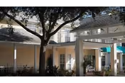 Exterior view of Bayou Gardens Assisted Living facility showing a covered entrance with white pillars, a large tree in the courtyard, and part of the two-story building with windows and a balcony.