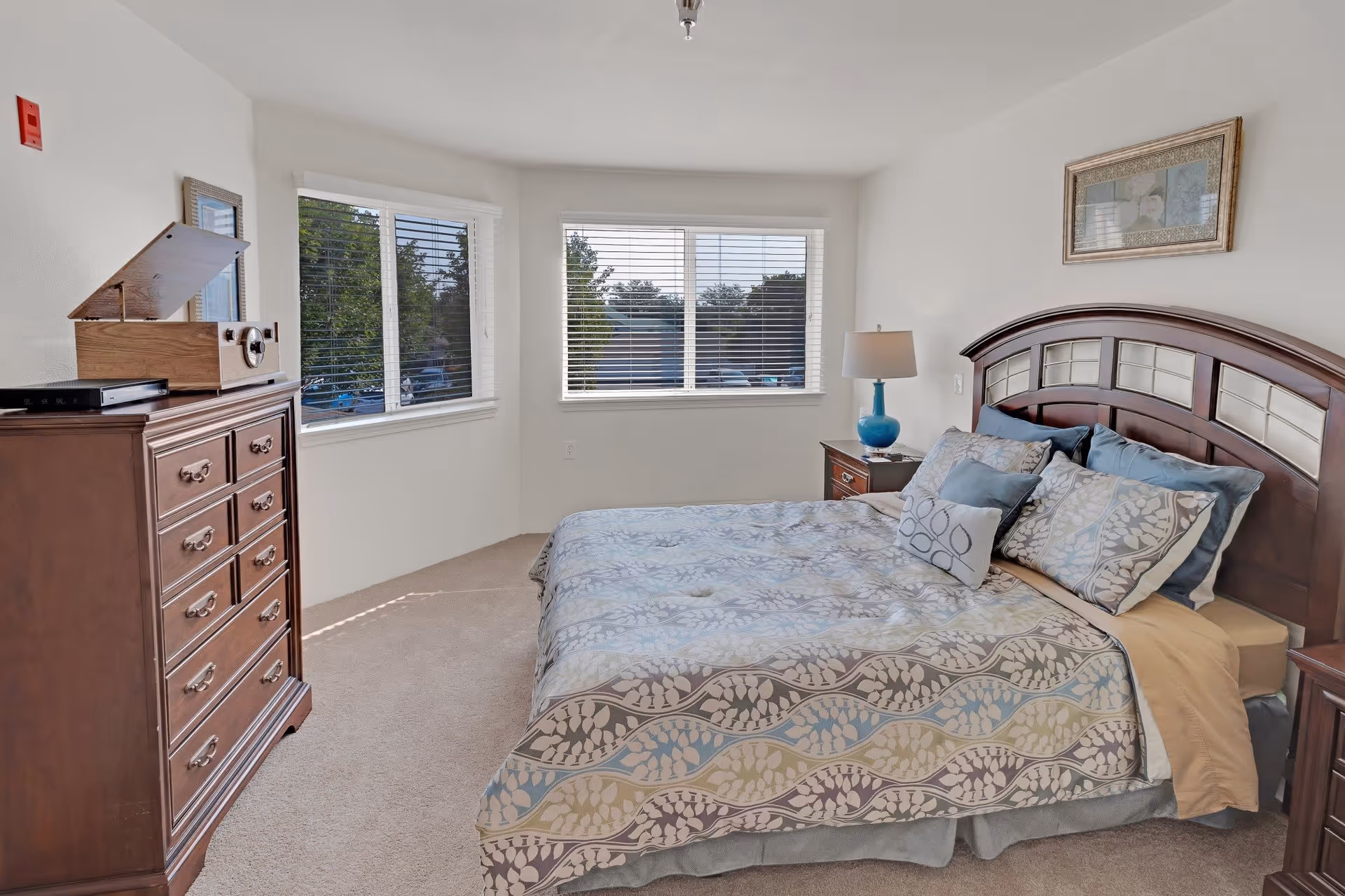 A bright bedroom with a large bed covered in patterned bedding and multiple pillows. There is a wooden headboard, a nightstand with a blue lamp, a wooden dresser with a record player on top, and two windows with white blinds letting in natural light.