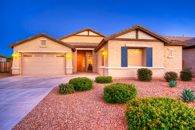 Exterior view of a single-story house with a two-car garage, beige stucco walls, blue shutters, and a landscaped front yard with bushes and gravel. The house is illuminated by warm outdoor lights at dusk.