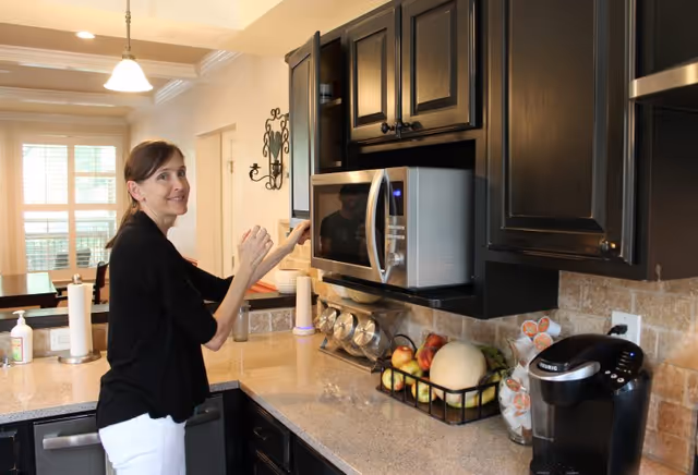 A woman standing in a kitchen area, reaching towards a microwave oven mounted above the countertop. The kitchen has dark cabinets, a Keurig coffee maker, a basket of fruit including apples and a melon, and a paper towel holder. The background shows a dining area with a table and chairs near windows with blinds.