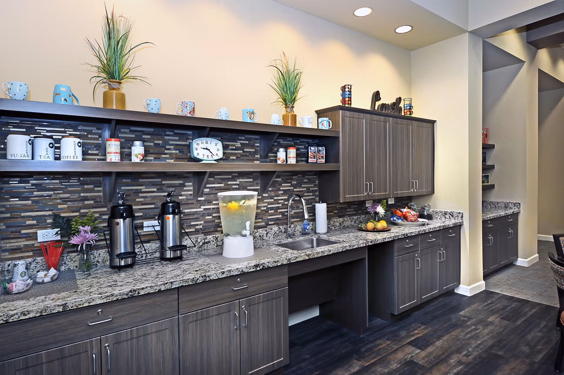 A modern kitchen area with dark wood cabinets and granite countertops. The backsplash features a mosaic tile design. On the counter, there are two coffee dispensers, a water dispenser with lemon slices, a sink, a plate with fruit, and various snacks. Shelves above hold decorative mugs, plants, and containers labeled sugar, tea, and coffee. The floor is dark wood, and the space is well-lit with recessed ceiling lights.