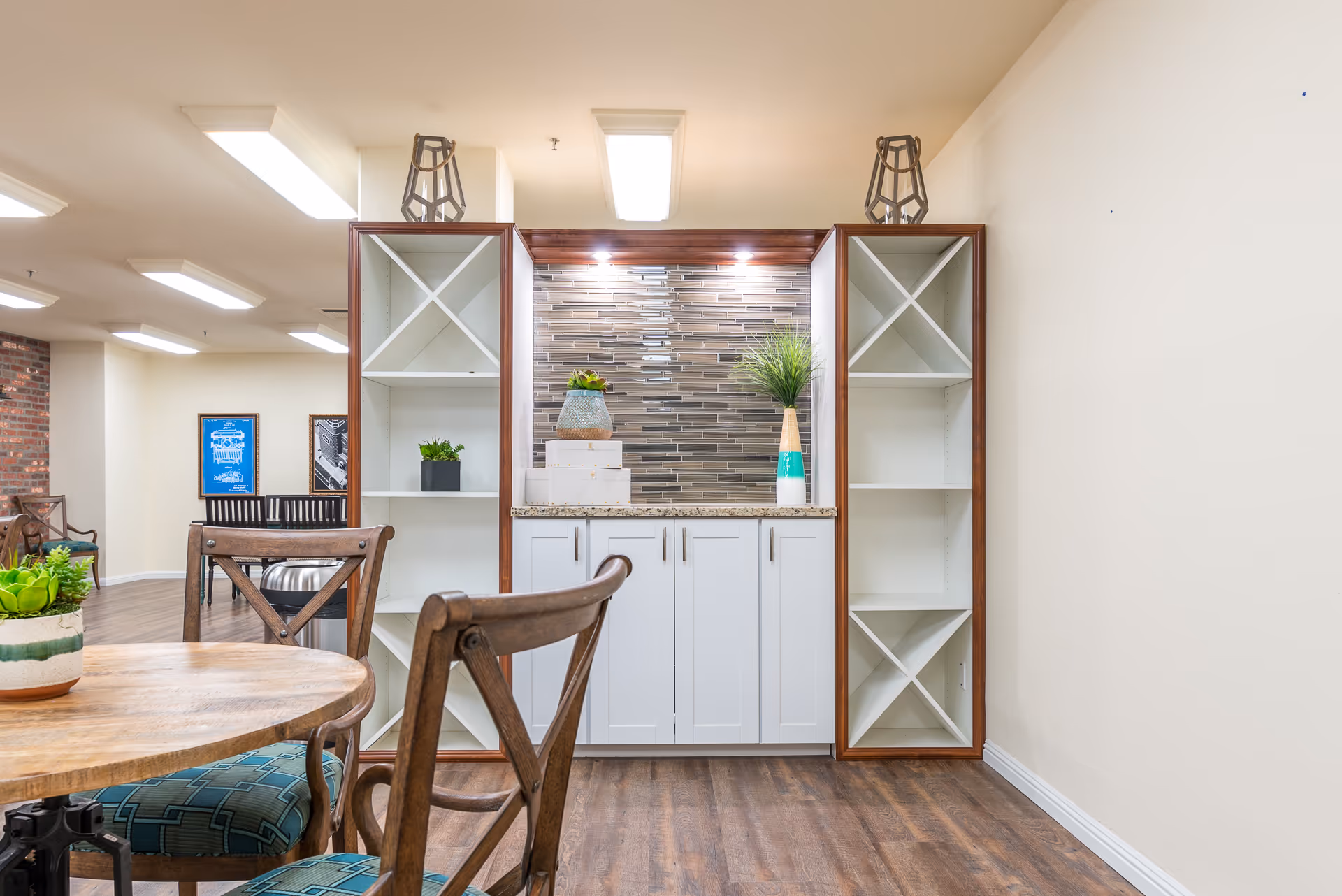 Interior view of a senior living facility common area with wooden chairs and a round table in the foreground. A built-in cabinet with white shelves and a granite countertop is centered against the wall, decorated with small plants and decorative boxes. The floor is wooden, and the ceiling has fluorescent lights.