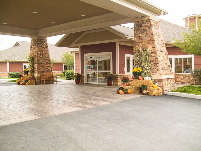 Entrance of a senior living facility with a covered drop-off area supported by stone pillars. The building exterior is red with stone accents and there are autumn decorations including pumpkins, hay bales, and potted plants near the entrance.