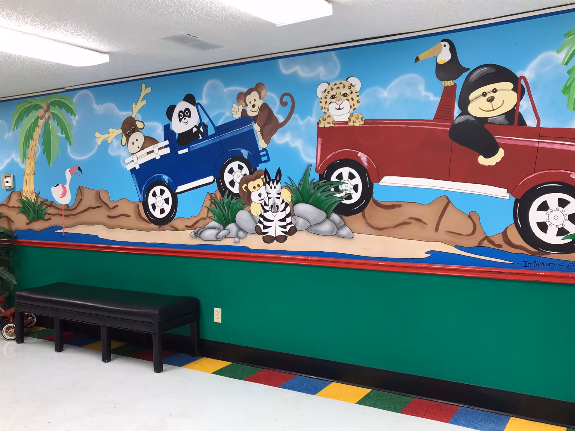 A colorful mural on a wall depicting cartoon animals including a moose, panda, monkey, leopard, toucan, flamingo, lion, and zebra riding in blue and red trucks. The mural is set against a blue sky with clouds and a desert-like landscape with rocks and plants. Below the mural is a green wall with a black bench and a multicolored tiled floor.