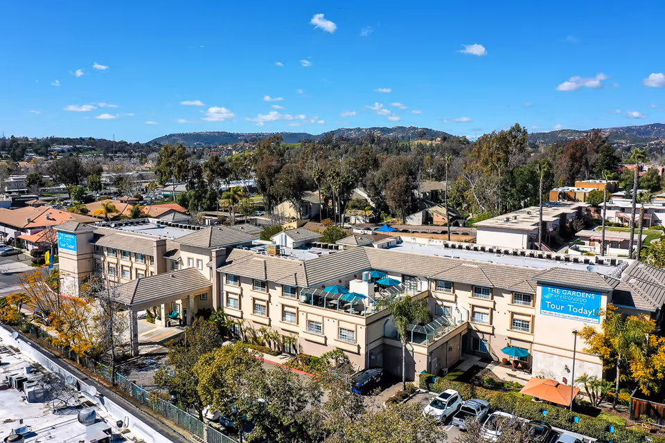 Aerial view of The Gardens at Escondido senior living building with patios, parking lot, and surrounding trees and hills under a blue sky.
