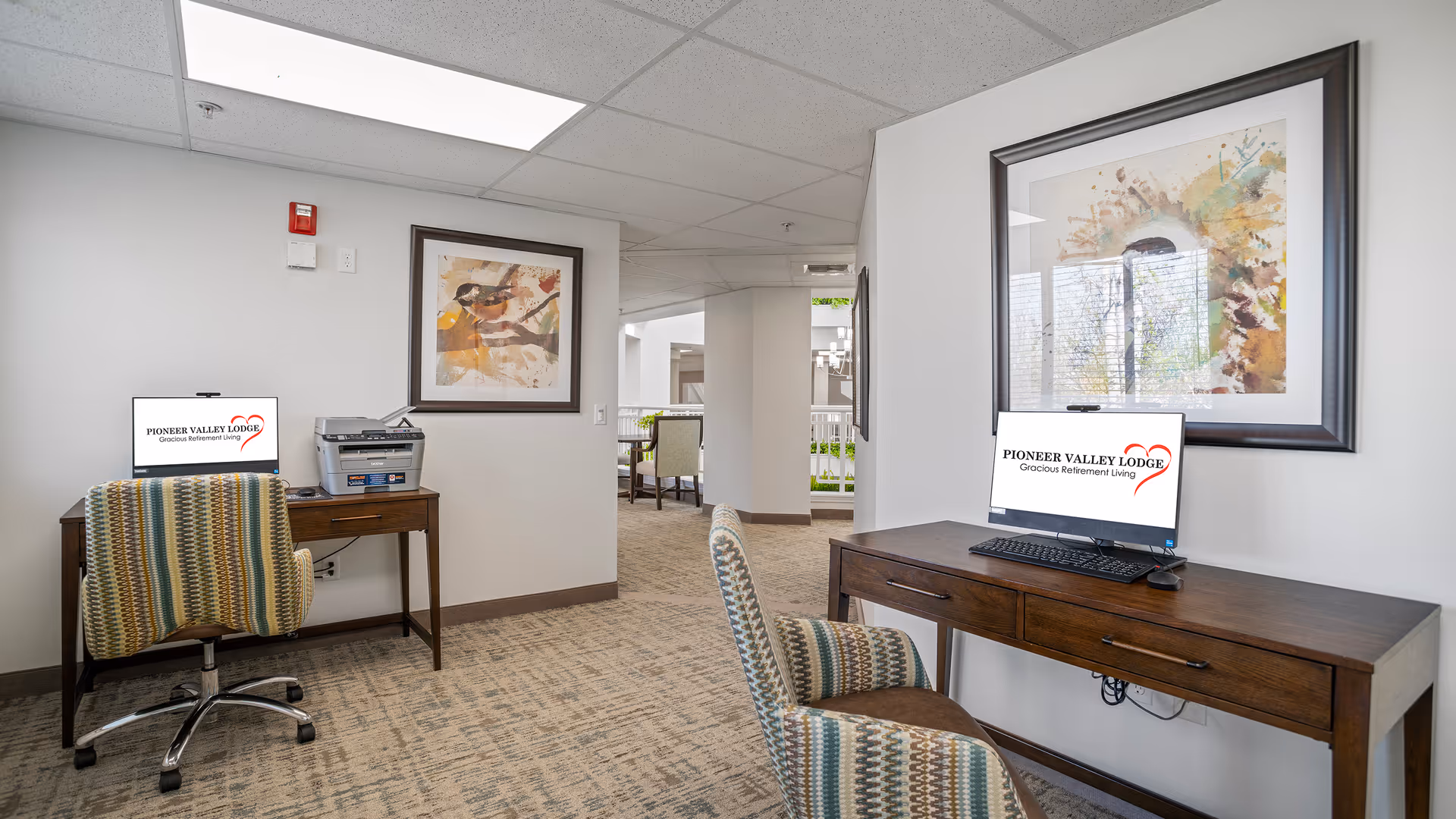 Two computer workstations with monitors and patterned chairs in a carpeted interior common area of a retirement facility.