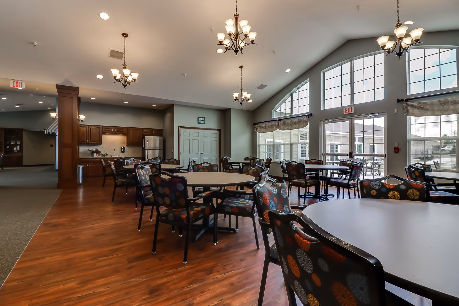 A spacious dining area with round tables and chairs featuring colorful patterned upholstery. The room has large arched windows allowing natural light to fill the space. There are chandeliers hanging from the ceiling and a kitchen area with wooden cabinets and stainless steel appliances in the background.