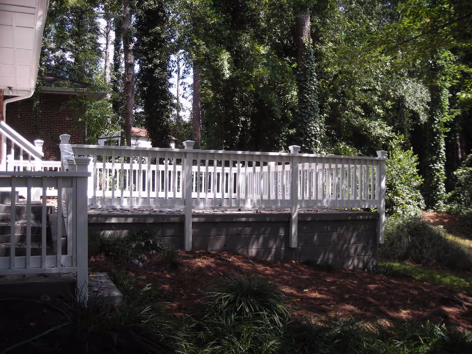 Outdoor view of a white wooden deck railing attached to a building, surrounded by tall trees and greenery with sunlight filtering through the leaves.