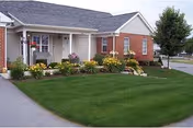 Exterior view of a single-story residential building with a front porch, well-maintained green lawn, and colorful flower beds along the front. A tree is visible on the right side near the sidewalk.